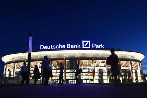 Mit einem neuen 73 Meter langen Schriftzug „Deutsche Bank Park“ präsentiert sich das beleuchtete Frankfurter Stadion im Abendlicht. Im Vordergrund befinden sich einige unscharfe Fans.